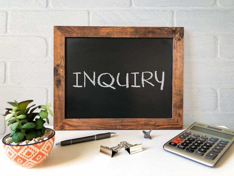 a blackboard on a counter with inquiry written on it. Also a plant in a red and white pot, a pen, a calculator, a top and 2 bulldog clips sit on the counter. The background is a whitewashed brick wall